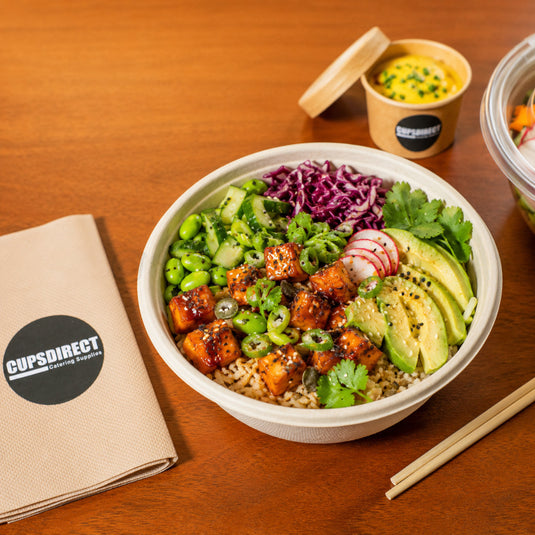 A prepared rice bowl with avocado, radishes, edamame, greens, purple cabbage, and glazed tofu sits on a wooden table. A branded napkin and small paper cup with sauce are visible, along with chopsticks and part of another container.